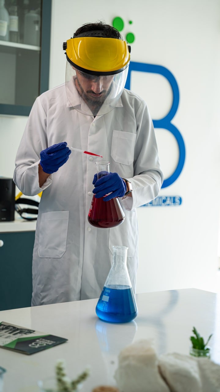 A chemist in a face shield adding drops to a flask of red solution