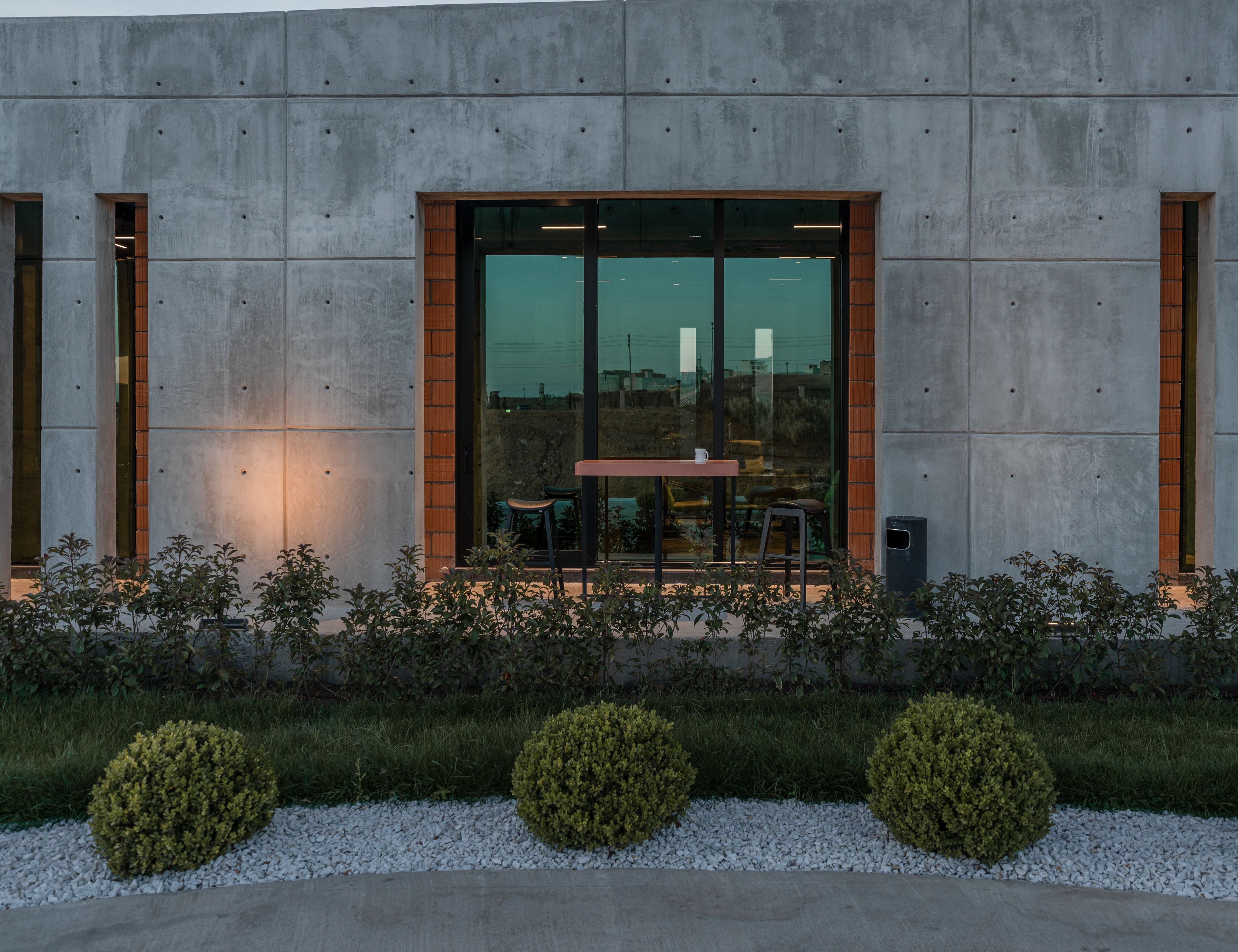 The head office's concrete facade at dusk, a lit window between brick reveals above clipped shrubs